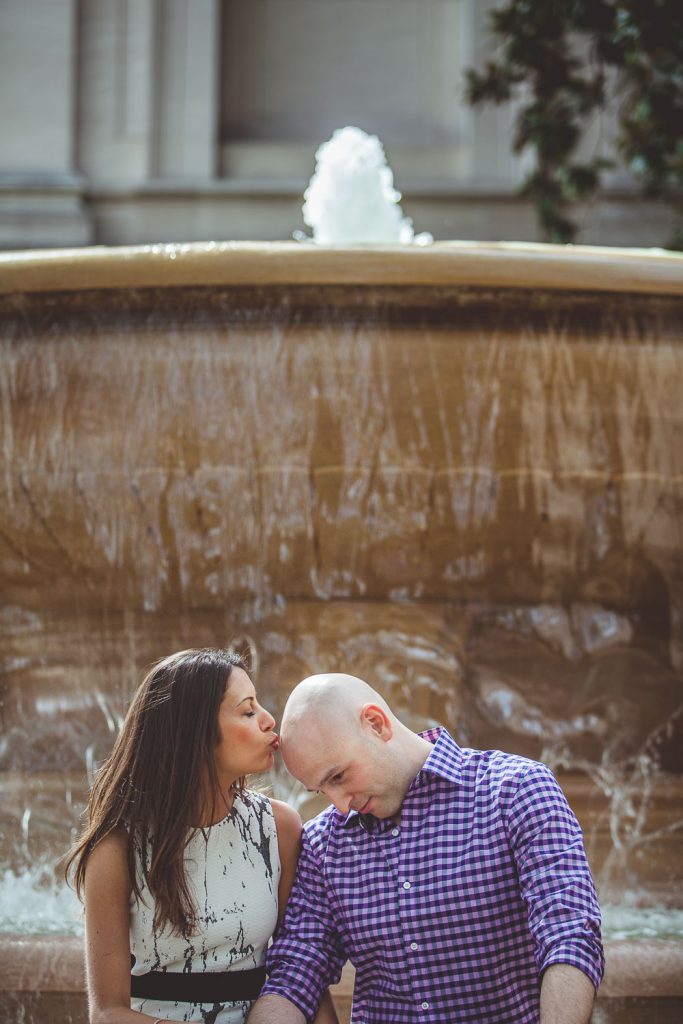 Black and white photo of an Engaged couple in front of a fountain in DC as she kisses his head.