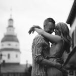 A black and white portrait of an engaged couple hugging in front of a downtown Annapolis building.