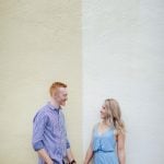 An engaged couple in Downtown Annapolis, holding hands in front of a yellow wall for their engagement photo portrait.