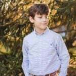 A boy in a blue shirt is posing for a portrait in front of a pine tree.