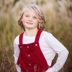 A little girl in red overalls standing in tall grass during sunset.