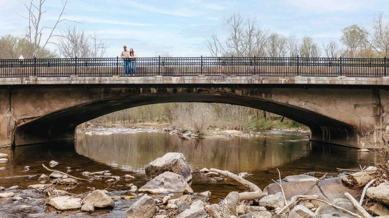 An engaged couple stands on a bridge in Old Ellicott City, capturing a romantic portrait against the scenic backdrop of the river.