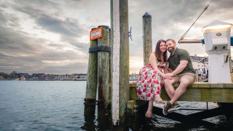 A couple sits on a dock with a boat in the background, capturing a beautiful candid portrait during their engagement photos session in downtown Annapolis.