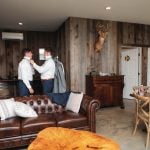 Two men, dressed in wedding attire, candidly putting on their ties in a living room at the Fleetwood Farm Winery.