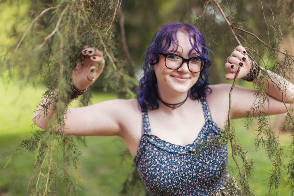 A high school senior with glasses and purple hair is leaning against a tree.