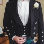 A groom in a kilt is preparing for his wedding, standing candidly in front of a mirror.