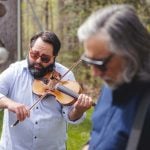 Two bearded men playing violin in a wooded area during a candid moment.