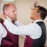 Two men preparing for a wedding while candidly adjusting each other's vests in a bedroom.