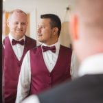 A man in a burgundy vest is candidly preparing for a wedding in front of a mirror.