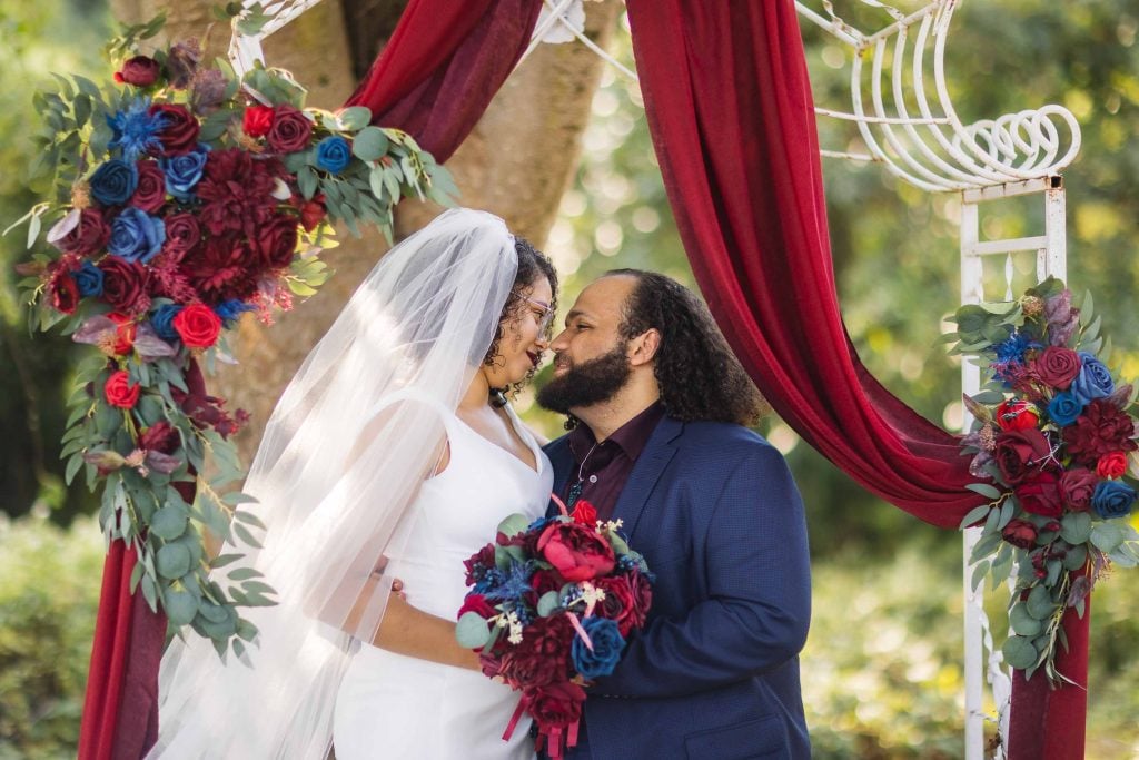 A bride and groom pose for a wedding portrait under a red and blue wedding arch.