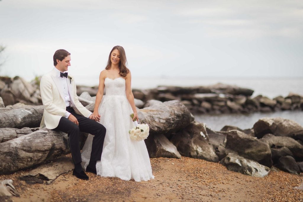 A wedding portrait of a bride and groom sitting on a log near the water.