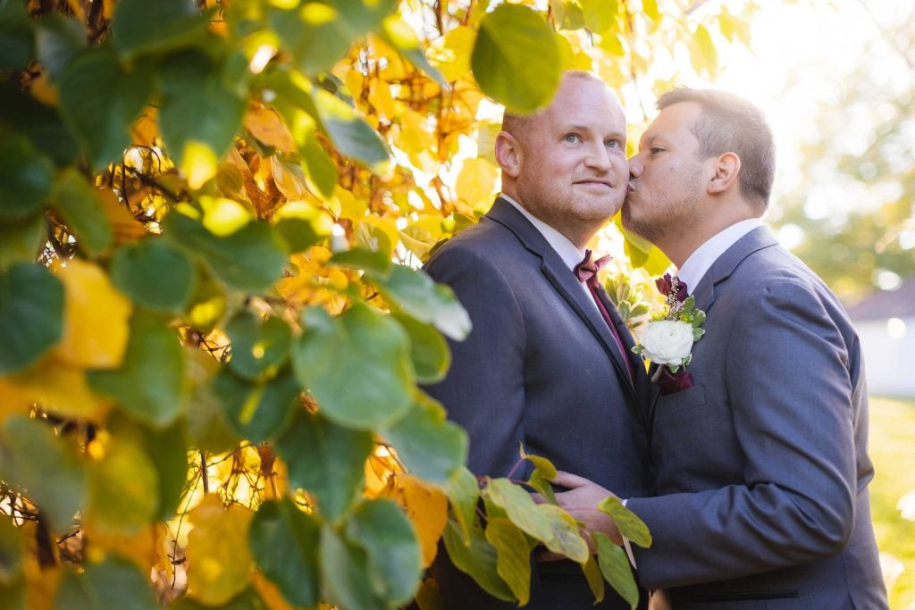 Two grooms sharing a wedding kiss in front of a tree.