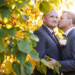 Two grooms sharing a wedding kiss in front of a tree.