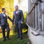 Two grooms holding hands in front of a fence, capturing their wedding portrait.