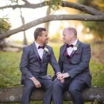 Two grooms sitting on a log in a wedding portrait.