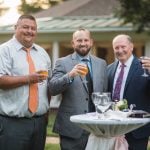 Three men in suits standing next to a table at a wedding reception with drinks.