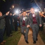 Two wedding grooms walking down a path with sparklers in their hands during the reception.