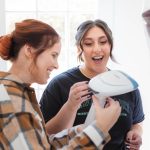 Two women candidly laughing while preparing for a wedding, using a hairdryer.