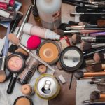 A pile of makeup brushes and cosmetics with wedding details on a table.