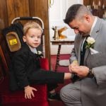 A candid man in a suit helping with wedding preparation by putting a boy's shoe on.