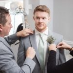 A candid man preparing a groom for the wedding by putting a boutonniere.