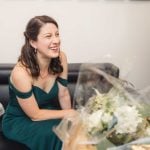 A woman in a green dress candidly laughs while sitting on a couch during wedding preparation.