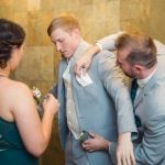 A man in a suit is candidly preparing another man for a wedding by helping him put on a boutonniere.