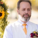 A man in a white vest and yellow tie is standing in front of sunflowers at a ceremony.