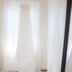 A wedding dress hangs in front of a mirror, showcasing intricate details.