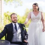 A candid wedding preparation with a bride in a wedding dress and a groom in a wheelchair.