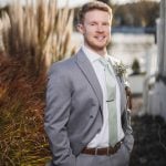 A groom in a wedding suit standing by a body of water.