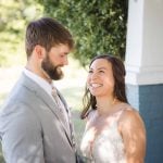 A candid wedding couple smiling at each other in front of a house.