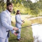 Two groomsmen posing for a wedding portrait, holding fishing rods in front of a pond.