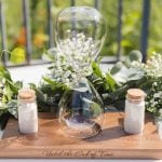 A glass hourglass on a wooden table with detailed flowers and greenery, perfect for a wedding.