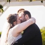 A bride and groom share a kiss during their wedding ceremony under a beautiful arch.