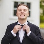 A man in a tuxedo adjusting his bow tie for a wedding portrait.