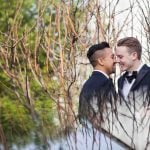 Wedding portrait of two men in tuxedos kissing in front of a tree.