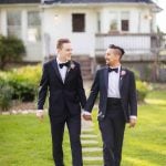 Two men in tuxedos holding hands in front of a house for a wedding portrait.