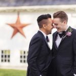 Two men in tuxedos are smiling in front of a barn for a wedding portrait.