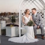 A wedding portrait of a bride and groom on a wooden bench by a dock.