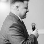 A man in a suit speaking at a wedding reception.