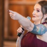 A woman with tattoos at a wedding reception posing near a microphone.