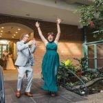 A bride and groom walk out of a building after their wedding ceremony with their hands in the air.