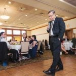 A man in a suit speaking into a microphone at a wedding reception.