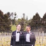 Two grooms in tuxedos standing in a vineyard for their wedding portrait.