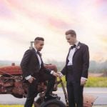 Two men in tuxedos posing next to a tractor at a wedding.