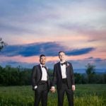 Two grooms in tuxedos standing in a field at sunset for their wedding portrait.