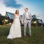 A wedding portrait of a bride and groom standing in front of a greenhouse at dusk.