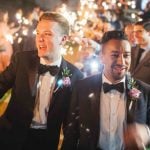 Two men in tuxedos holding sparklers at a wedding reception.