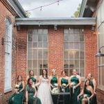 A group of bridesmaids in emerald dresses pose in front of a Savage Mill building.